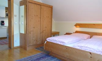 A cozy bedroom with a double bed and a wooden wardrobe. The walls are bright and the floor has a warm wood tone.