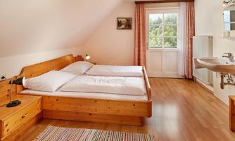 A cozy bedroom with two wooden single beds and a window that overlooks the green landscape. The floor is wooden, and a carpet lies in front of the beds.