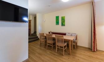 A cozy dining area with a wooden table and chairs. The walls are light, and there are some green pictures as well as a television on the wall.