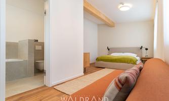 A modern hotel room with a double bed and a cozy seating area. The room has a clean, minimalist style and plenty of daylight.
