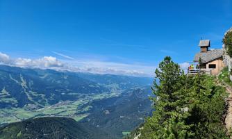 A glorious mountain landscape with green valleys and a clear blue sky. In the foreground stands a small mountain hut, surrounded by trees.