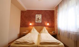 A simple bedroom with two single beds and a warm, rustic color tone. Bathed in light from a large window with curtains.