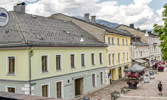 A picturesque street with cozy houses in soft colors. In the foreground stands the Schachner guesthouse, and there are cafés as well as trees that create an inviting atmosphere.