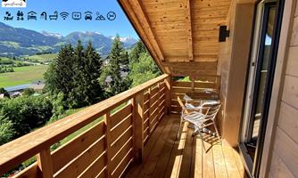 A cozy balcony with wood paneling, two chairs and a table. In the background, green meadows and mountains are visible.