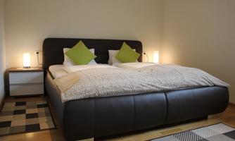 A modern bedroom with a large double bed and two bedside tables. The decoration consists of green cushions and warm light.