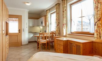 A bright and cozy room with a kitchenette, a dining table, and wooden shelves. Large windows provide plenty of daylight.