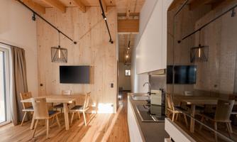 A modern kitchen with a dining area featuring wooden chairs and a table. The room has a bright, cozy atmosphere with wooden paneling and large windows.