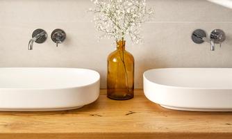 A modern bathroom with two white sinks on a wooden shelf. A vase with fresh flowers is placed in the center, and towels are neatly arranged.