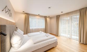 A bright and modern bedroom with hardwood flooring. The room features a large bed and has windows with curtains that let in plenty of daylight.