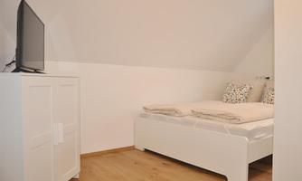 A cozy bedroom with a white bed and decorative cushions. Next to the bed, there is a television on a white cabinet.