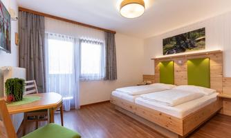 A bright and modern room with two beds and wooden decor. There is a table with chairs and large windows that let in plenty of daylight.