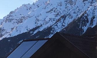 A snow-covered mountain in the background with a blue sky and some clouds. In the foreground, the roof of a house is visible.