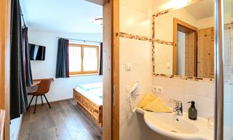 A modern bathroom with a sink, mirror, and shower. In the background, a cozy bedroom with a window and a television is visible.