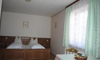 A simple room with a double bed and two pillows. In the background, there is a table and a window with curtains.