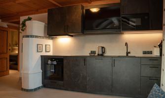 A modern kitchen with dark cabinets and a stainless steel sink. A television is mounted on the wall, and there is a kettle as well as a coffee machine.