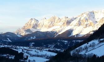A beautiful mountain landscape with snow-covered peaks and a clear blue sky. In the valley, green meadows and small villages stretch out.