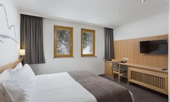 A modern hotel room with a large bed and wooden details. The windows offer a view of the snow-covered landscape.