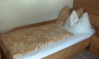 A simple bed with a floral blanket and two pillows. The room has a wood-paneled wall and a colorful rug.