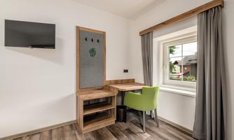 A modern room with a desk and a chair in green. A television hangs on the wall and a window lets in daylight.