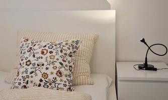 A cozy bedroom with a white bed and a colorful pillow. Next to the bed is a simple nightstand with a small lamp.