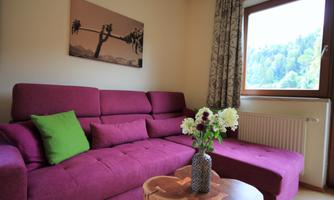 A cozy living room with a purple couch and a small wooden table. Through the window, you can see a green, wooded landscape.