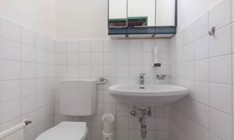 A small bathroom with white tiles. It contains a sink, a toilet, and a mirror above the sink.