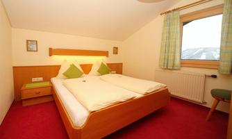 A cozy bedroom with two single beds and light wooden furniture. It has a red carpet and a window with a view outside.