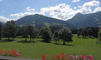A picturesque view of green meadows and large trees, surrounded by mountains. The sky is clear and some clouds are visible.