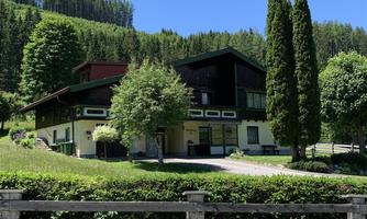 A modern house surrounded by green trees and a well-maintained garden. Gentle hills can be seen in the background.