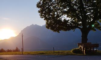A picturesque sunset over the mountains. A large tree stands beside a bench on a meadow.