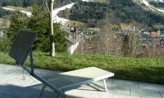 A lounger is located on a terrace overlooking a picturesque landscape. In the background, green hills and snow-capped mountains can be seen.
