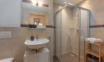 A modern bathroom with a shower, a sink, and a shelf. The walls are light tiled and there are towels and toiletries.