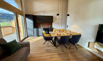 A modern dining room with a large table and colorful chairs. Plenty of daylight streams through the windows, and there is a cozy seating area.