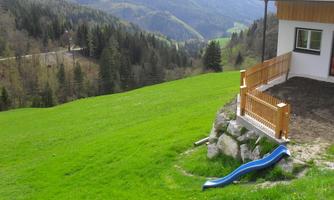 A green, hilly landscape with a house and a slide in the foreground. In the background, mountains and a clear sky can be seen.