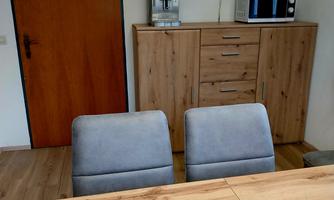 A modern room with a wooden table and two gray chairs. On the wall, there is a television as well as a kitchenette with a coffee machine and microwave.