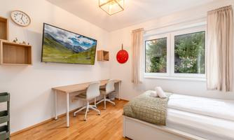 A modern bedroom with a large window and a TV on the wall. The room has bright furniture, a desk, and a comfortable bed.