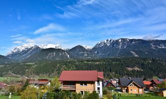 Mountain landscape with snow-capped peaks and a green valley. Small houses at the foot of the mountains under a clear sky.