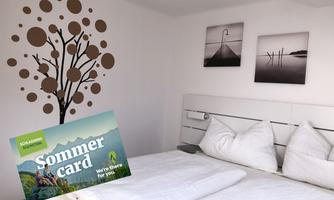 A modern bedroom with a stylish tree motif on the wall and atmospheric black-and-white photos. The summer map is lying on the bed and invites to activities.
