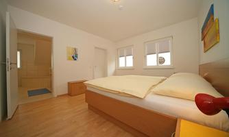 A bright bedroom with a large bed and two windows. The floor is made of wood and there is a small nightstand next to the bed.