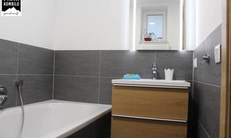 A modern bathroom with a bathtub and a wooden dresser. The walls are in shades of gray and white, and there is a large mirror with lighting.