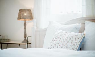 A cozy bedroom with a bed, white pillows, and a decorative pillow. Next to the bed, a stylish lamp stands on a small table.
