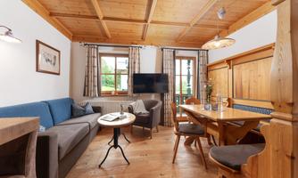 A cozy living space with a wooden ceiling, a blue sofa, and a dining table. There are large windows that let in plenty of light.