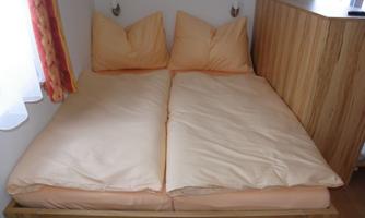 A cozy double bed with beige pillows and a wooden frame. The wall is light and there is a window with curtains.