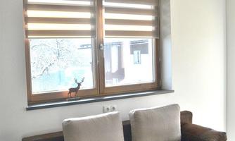 A modern living room with a brown sofa and two cushions. Above the sofa hangs a stylish lamp, and a window offers a view outside.