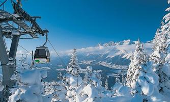 A cable car travels through a snow-covered landscape. In the background, majestic mountains and a clear blue sky can be seen.