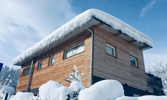 A modern wooden house with a snow-covered roof. The garden is also covered by a thick layer of snow.