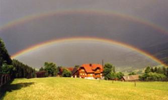 A beautiful rainbow spans a rural scene with a red house and green meadows. The sky is overcast, giving the landscape a dramatic mood.