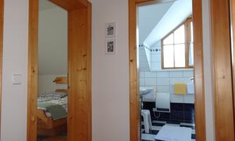 A bright hallway with wooden frames and two open doors. On the left is a bedroom and on the right is a bathroom.