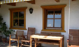 A cozy outdoor area with wooden chairs and a table. In the background, there are windows with yellow frames and a plant in a pot.