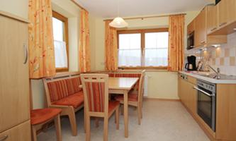 A bright kitchen with wooden furniture and a dining table. The windows are decorated with orange curtains.
